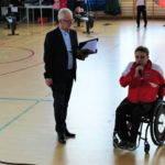zdjęcie konferansjera, obok trener szermierki na wózku inwalidzkim mówiący przez mikrofon