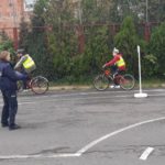 Dwójka dzieci na rowerach w kamizelkach odblaskowych i kaskach jedzie na rowerach pod okiem policjantki