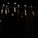zdjęcie wykonanie w ciemności, z ciemności wyłaniają się postaci trzymające na patykach papierowe statki, z lewej kobieta owinięta w biało-szare szaty dmuchająca na papierowe statki