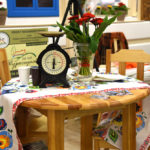 stół z dwoma krzesłami, na stole obrus, wazon z kwiatami oraz waga